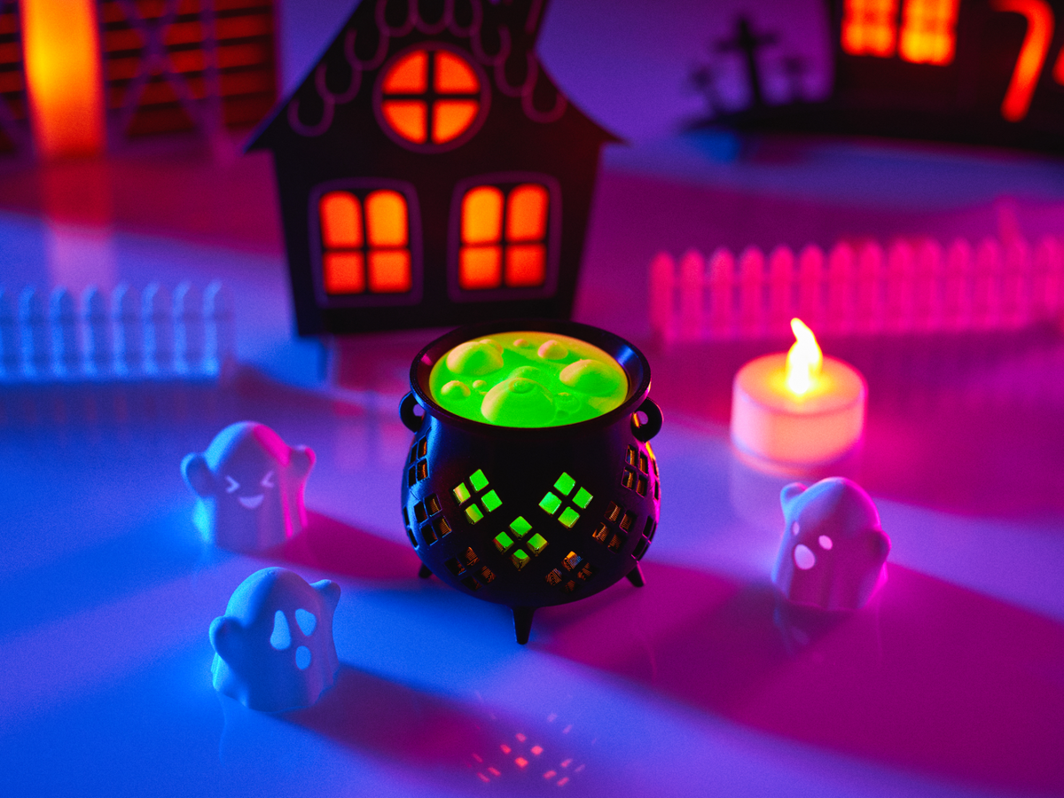 Halloween Cauldron Tealight Holder — black cauldron with green glowing top and diamond lattice light pattern, surrounded by ghost figurines and haunted house scene