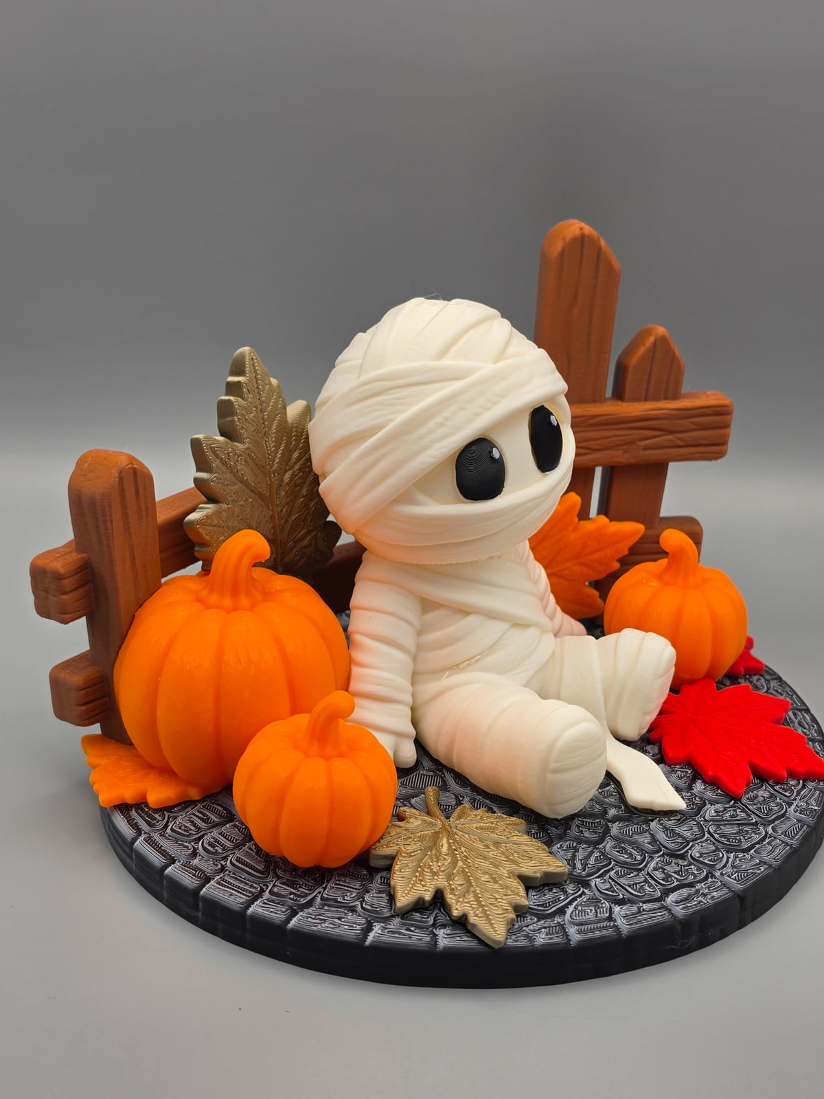 Halloween Mummy Sitting Figure — side-left angle showing wooden fence, gold autumn leaf and pumpkin arrangement