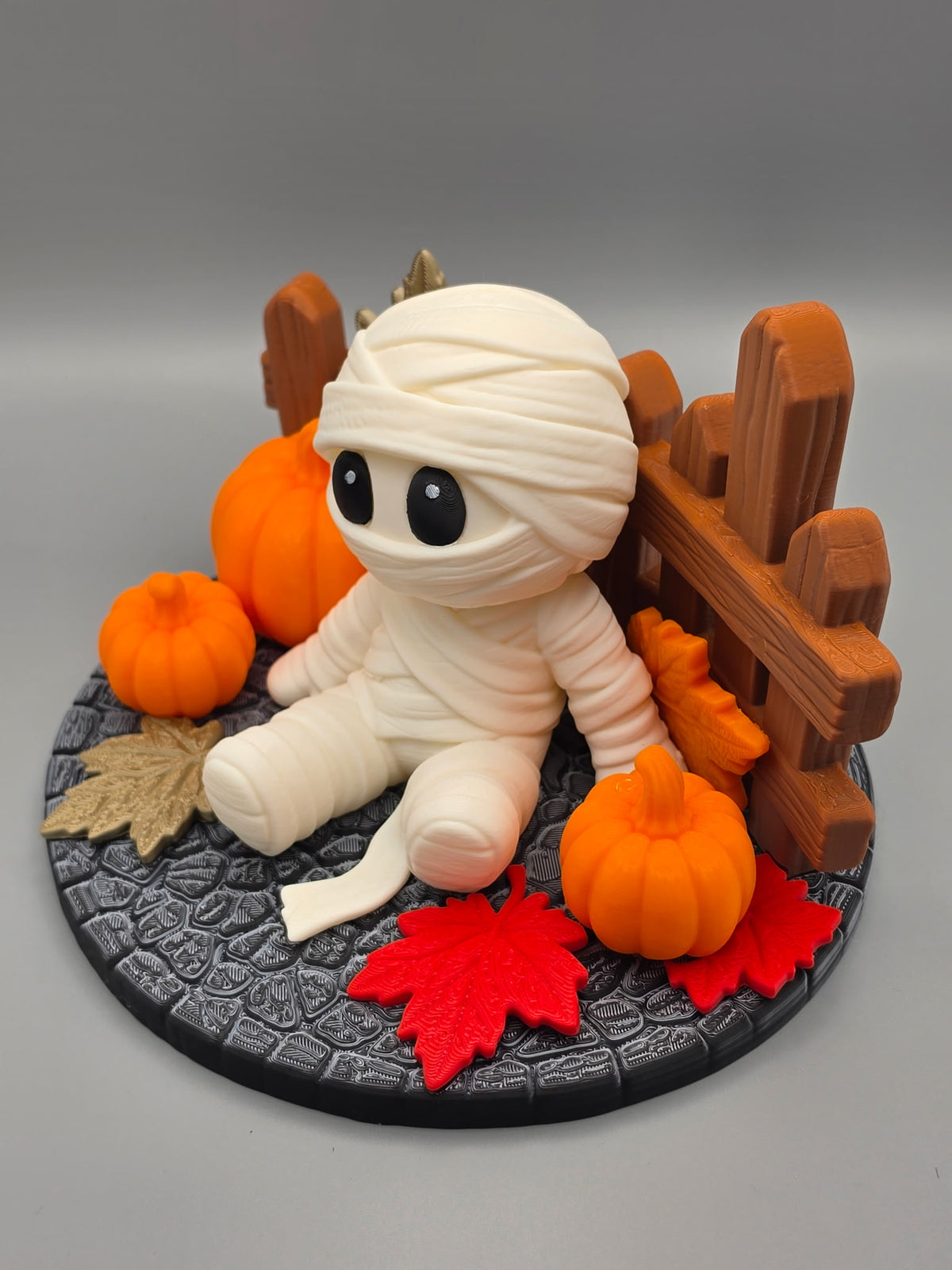 Halloween Mummy Sitting Figure — three-quarter right angle showing full scene with pumpkins, red leaves and fence detail