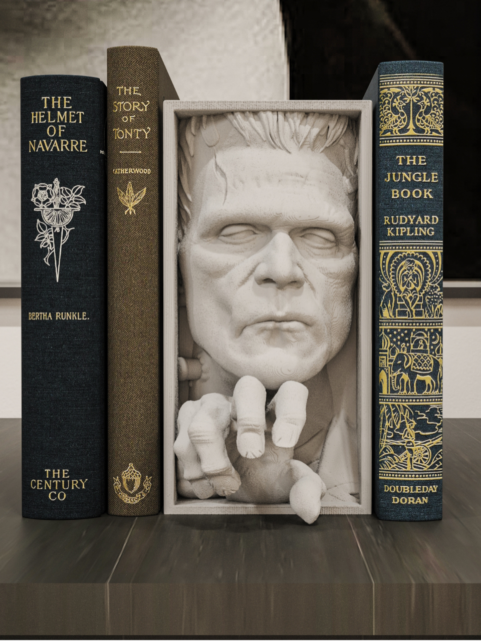 Frankenstein monster relief sculpture grey framed box used as book nook displayed between vintage hardcover books The Helmet of Navarre and The Jungle Book on a dark wooden shelf gothic library bookshelf styling the monster face and reaching clawed hand visible between book spines atmospheric moody editorial