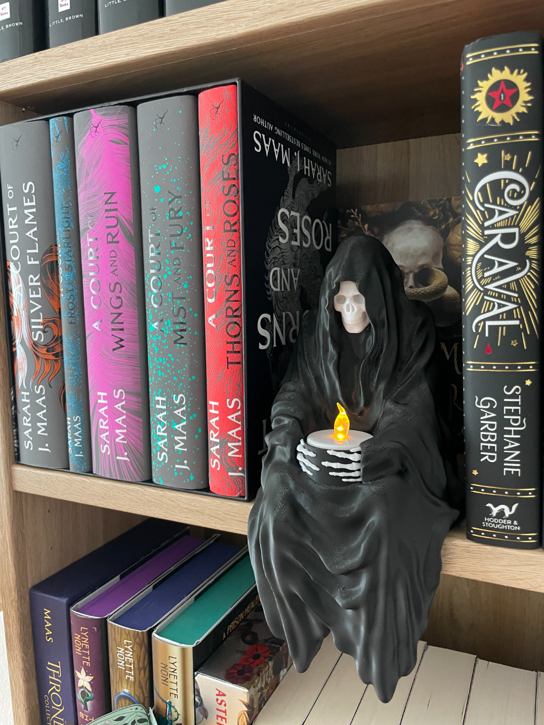 Grim Reaper shelf sitter figurine black matte hooded skull face flowing dark robes extraordinary fabric fold complexity skeletal hands holding glowing LED candle seated between colourful fantasy hardcover books on wooden bookshelf lifestyle hero product shot