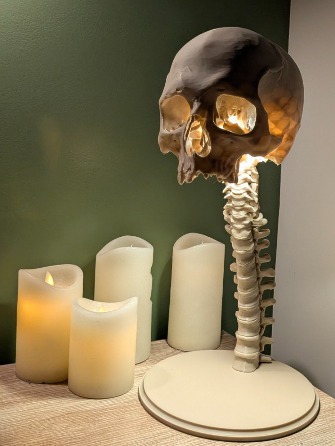 Skull and Spine Tealight Lamp — three-quarter front view showing amber eye socket glow with candles in background on wooden shelf against dark green wall