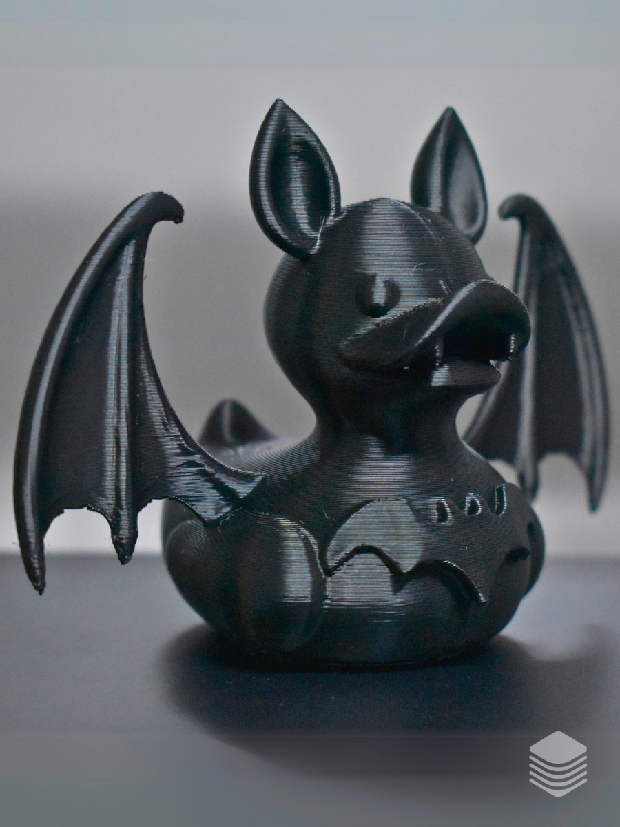 Gothic Bat Duck Sculpture — front three-quarter view: matte black rounded duck body, two large upright pointed bat ears at crown, broad flat duck bill slightly open, two large bat membrane wings with vein lines extending both sides, defined collar ring at neck, three circular chest studs, small rounded tail nub visible at rear