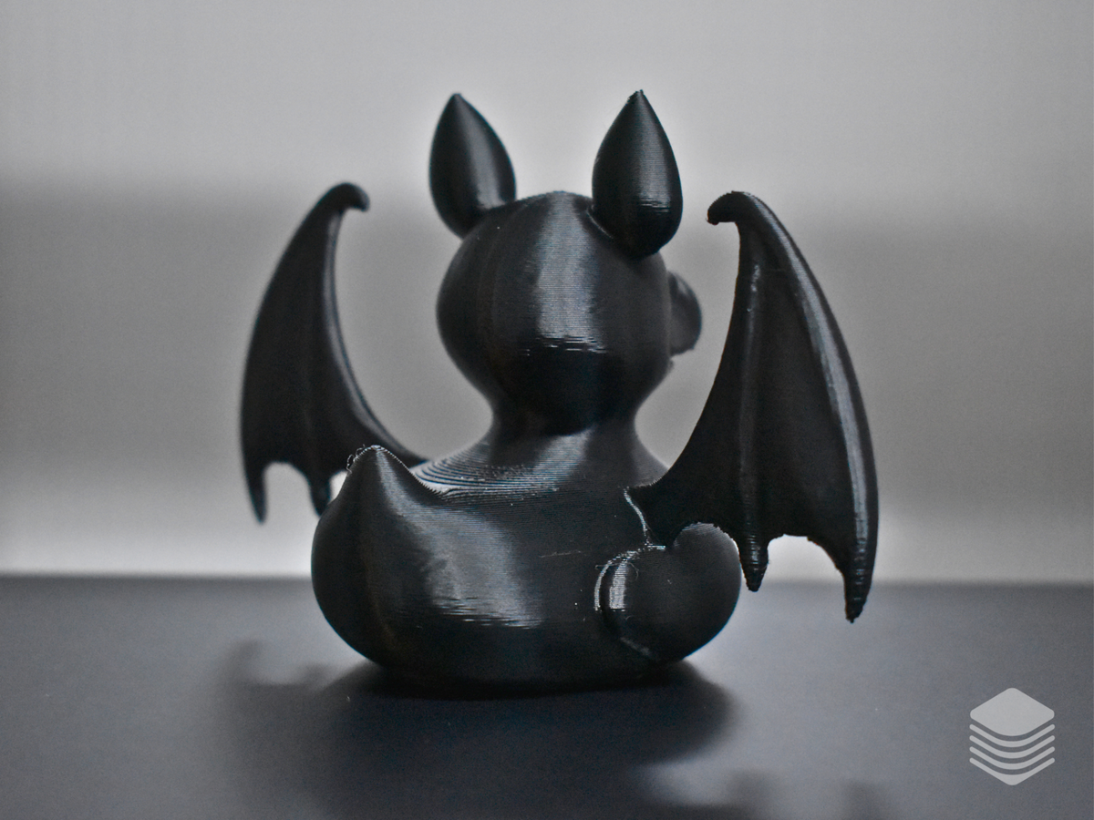 Gothic Bat Duck Sculpture — rear view: matte black rounded duck body from rear, two upright pointed bat ears from behind, bat wings extending both sides with membrane vein lines, small rounded tail nub at rear base, collar ring visible from rear