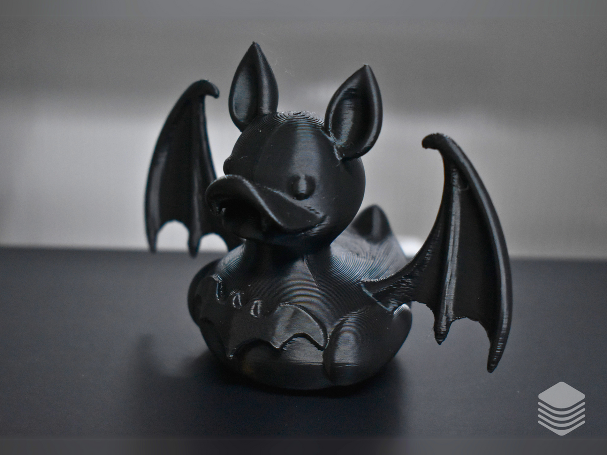 Gothic Bat Duck Sculpture — left front three-quarter view: matte black duck body, bat ear forms at crown, broad duck bill from left angle, left bat wing membrane vein detail prominent, collar ring and chest studs visible, stable flat base