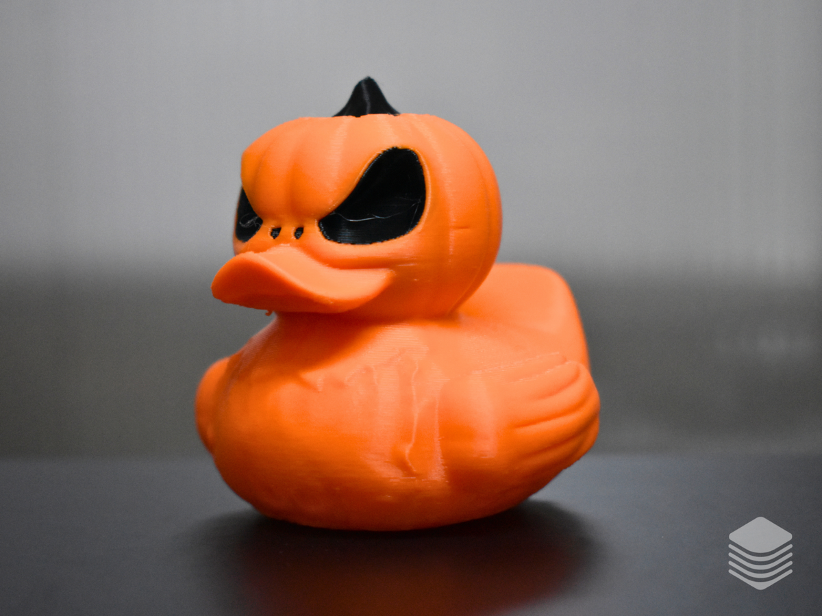 Halloween Pumpkin Duck — left three-quarter view: vivid orange pumpkin head and body with vertical lobe divisions from left angle, angry carved face with single black eye aperture and brow ridge visible, black stem at crown, orange duck bill from left, body lobe lines and wing form