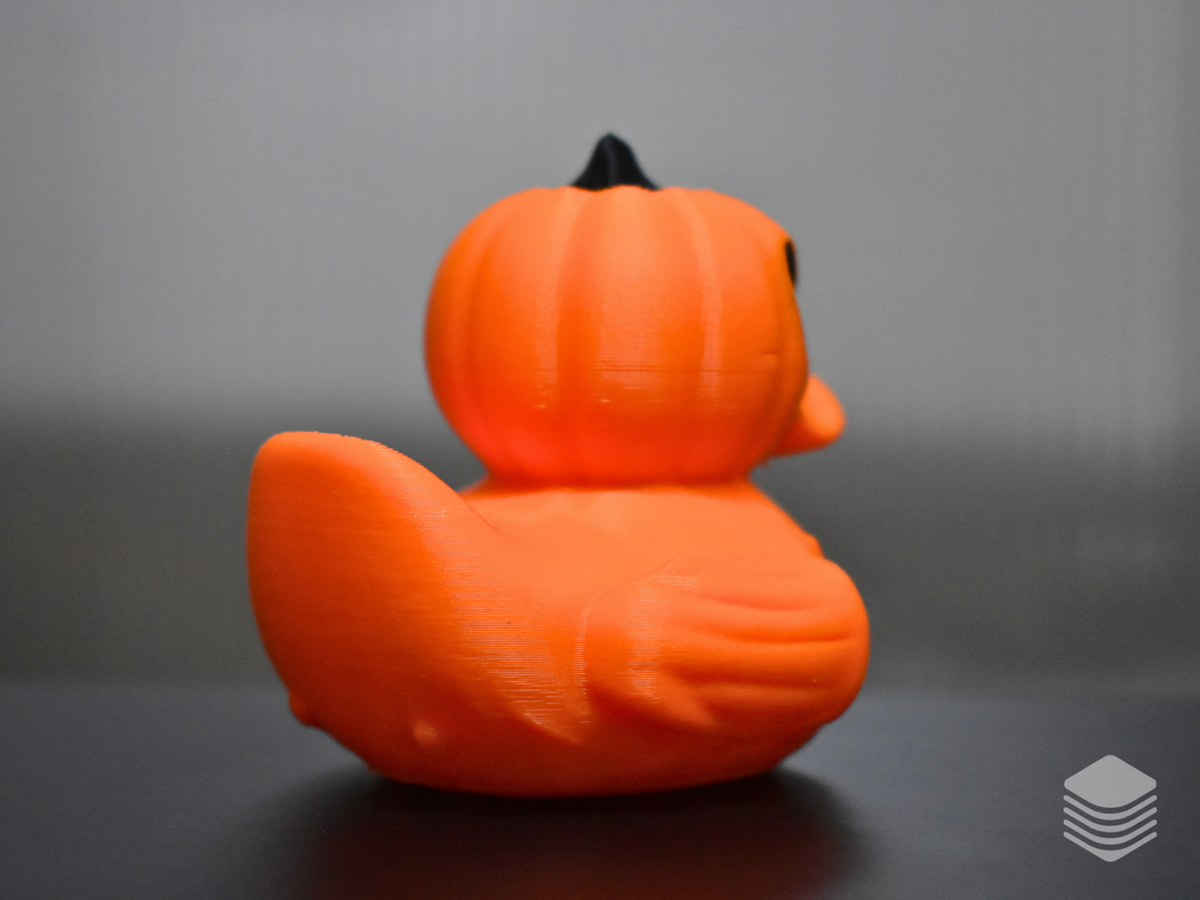 Halloween Pumpkin Duck — left rear three-quarter: vivid orange pumpkin body and head from rear left angle, vertical lobe divisions across full body and head, black stem at crown tip, orange tail form, wing form with lobe detail