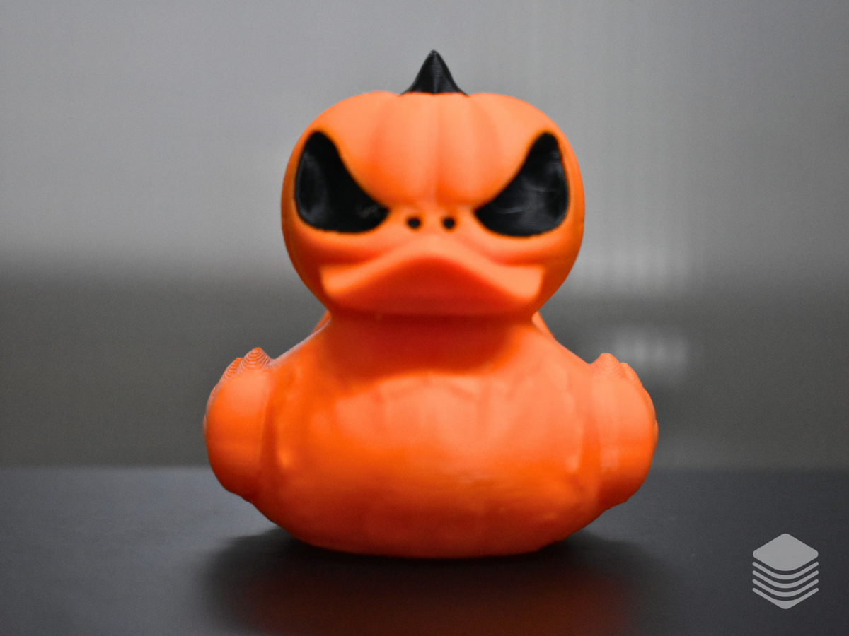 Halloween Pumpkin Duck — full front face-on: vivid orange pumpkin duck head-on, two large angled black void eye apertures with heavy angular furrowed brow ridges, three black nostril punches, orange integrated duck bill, black pointed stem at crown, full vertical pumpkin lobe body visible at both sides