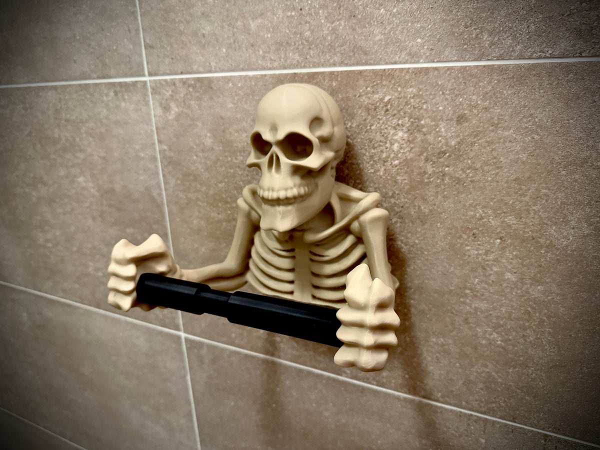 Skeleton toilet roll holder white bone colour full skeleton upper body emerging from wall holding black metal toilet roll bar between skeletal hands full ribcage detail anatomical skull face wall mounted bathroom hero product shot no roll