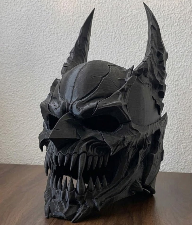Demon Skull Helmet Sculpture in deep charcoal obsidian — three-quarter front view on wooden surface showing full horn sweep, cracked plate dome, projecting brow ridge, deep eye sockets and wide open serrated fang jaw with jagged torn plate edges