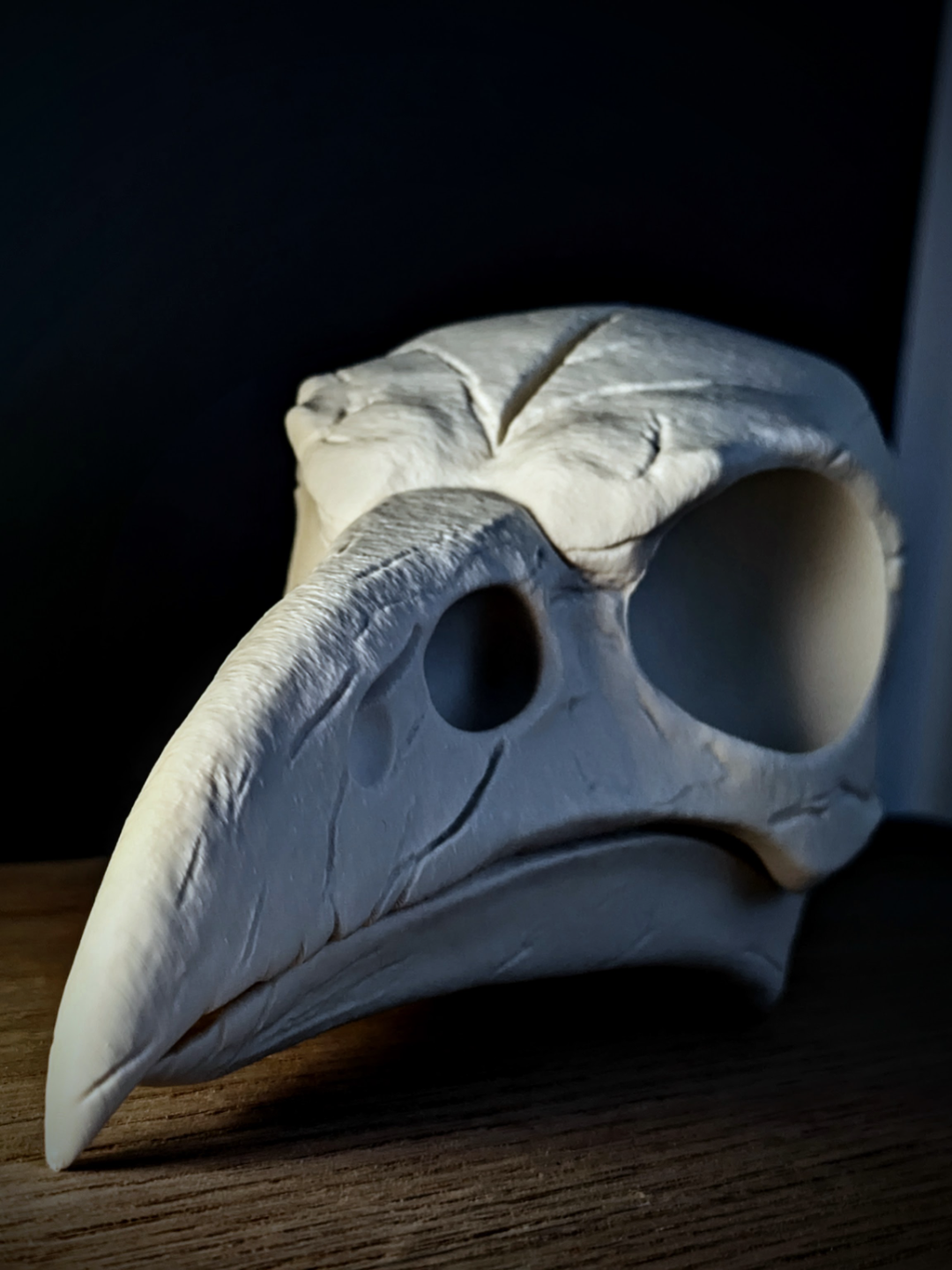Anatomical Raven Skull Sculpture in white — left side profile showing large primary orbital cavity, elongated curved beak, fenestration openings and cranial suture detail on dark oak surface