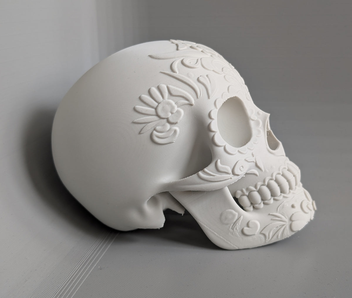 Day of the Dead Floral Skull Sculpture in white — side profile view showing full cranial floral relief, smooth rear occipital and open jaw structure on grey surface