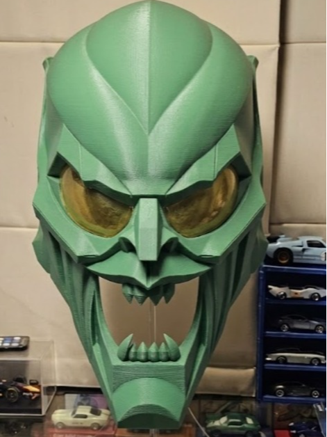 Geometric Villain Horror Mask Sculpture in green — front view showing faceted geometric cranial panels, prominent brow ridge, deep-set yellow lens eye cavities, angular cheekbones, open snarling jaw with lower fang row and swept temple guards