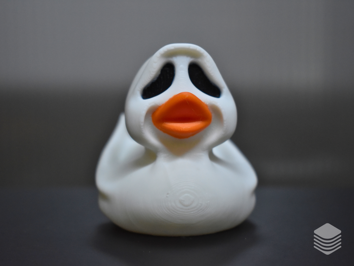 Halloween Ghost Duck Sculpture — full front face-on view: pure white ghost sheet with rounded hood peak, two large deeply recessed black teardrop void eye apertures, vivid orange duck bill at face centre, ghost sheet drape folds at both sides of face, flame-form sheet extensions visible at both rear sides