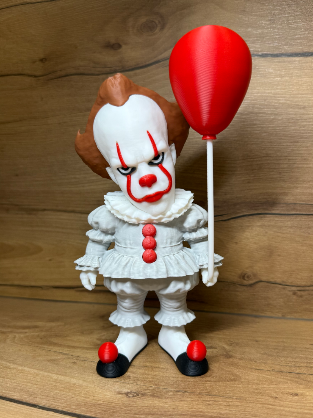 Pennywise IT Horror Minifigure Sculpture — front view showing white clown face with vivid red arched brow markings, yellow-gold eyes, red spherical nose dome, wide red smile lines, warm auburn tufted clown hair with bald crown dome, large white Elizabethan ruffled collar, white clown suit with three large red spherical pompom buttons, white ruffled waist skirt tier, right hand raised holding vivid red teardrop balloon on white stick, red pompom-tipped black shoes