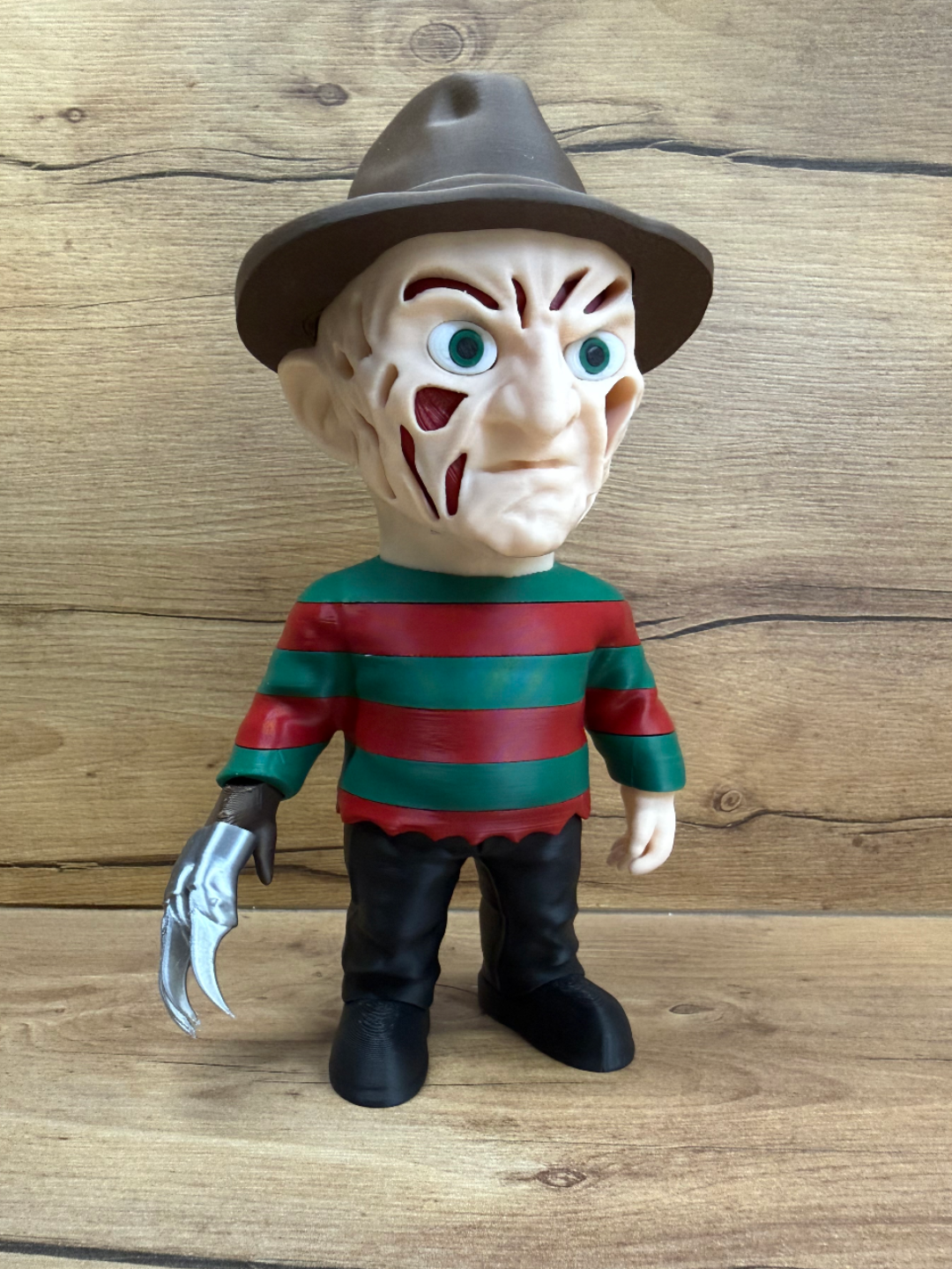Freddy Krueger Nightmare on Elm Street Minifigure Sculpture — front view showing dark olive-brown felt fedora with hat band, heavily scarred and burned face with organic red-brown scar tissue masses, open void apertures in scar tissue, wide teal-green eyes with white sclera, red and dark green alternating horizontal stripe sweater, bare left hand, right hand wearing four-blade silver razor glove with brown leather base, black trousers and black boots