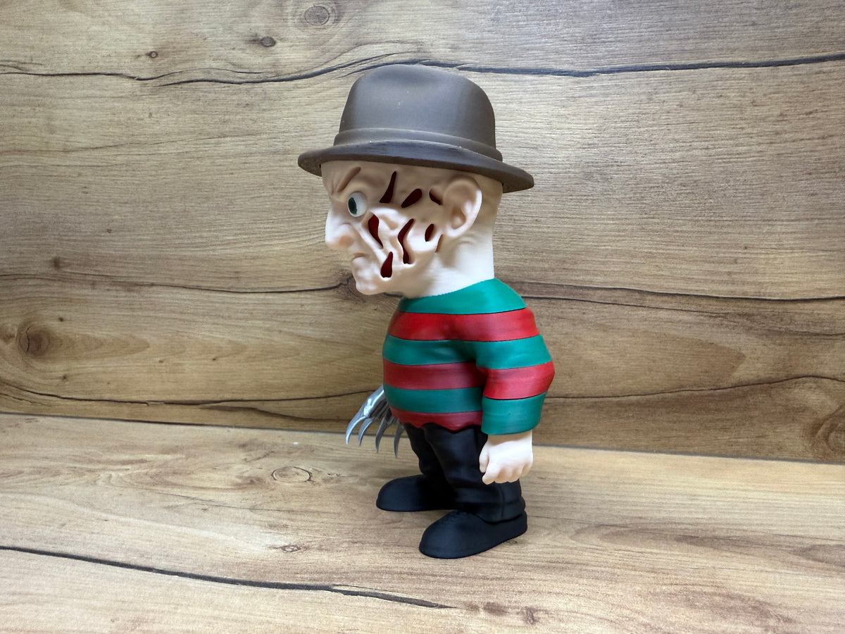 Freddy Krueger Nightmare on Elm Street Minifigure Sculpture — left side profile view showing felt fedora brim profile and dome crown, scarred face left profile with open void scar apertures and single teal-green eye visible, red and green stripe sweater left sleeve with individual stripe bands, bare left hand at side, right razor glove partially visible, black trouser leg and black boot profile