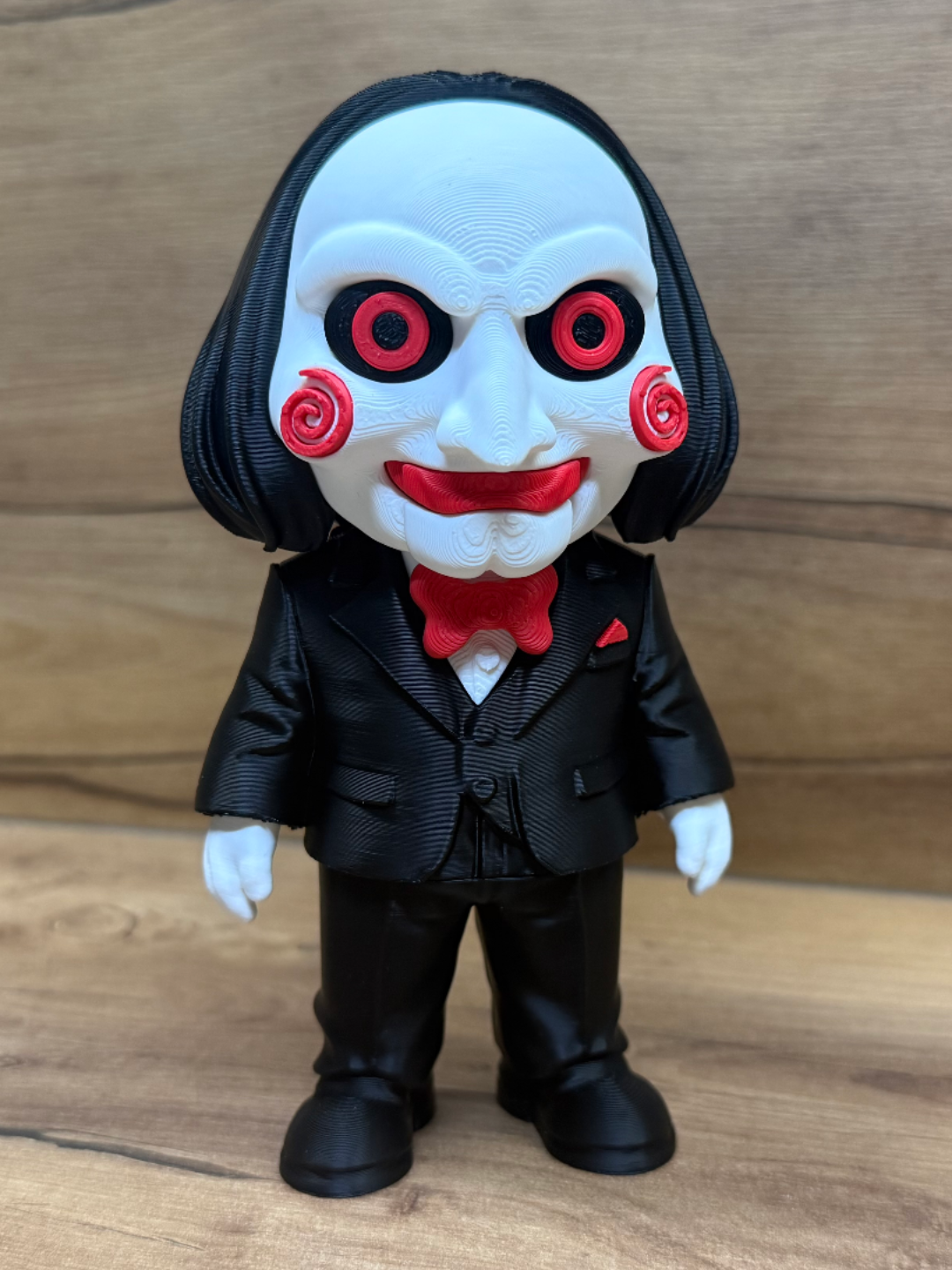 Billy the Puppet Saw Minifigure Sculpture — front view showing white ventriloquist doll face with deep forehead wrinkle lines, large red spiral cheek rosettes, red-ringed black eyes with centre pupils, wide open red-lipped grin with upper teeth, long black bob hair, black formal suit with red bow tie and pocket square and white-gloved hands