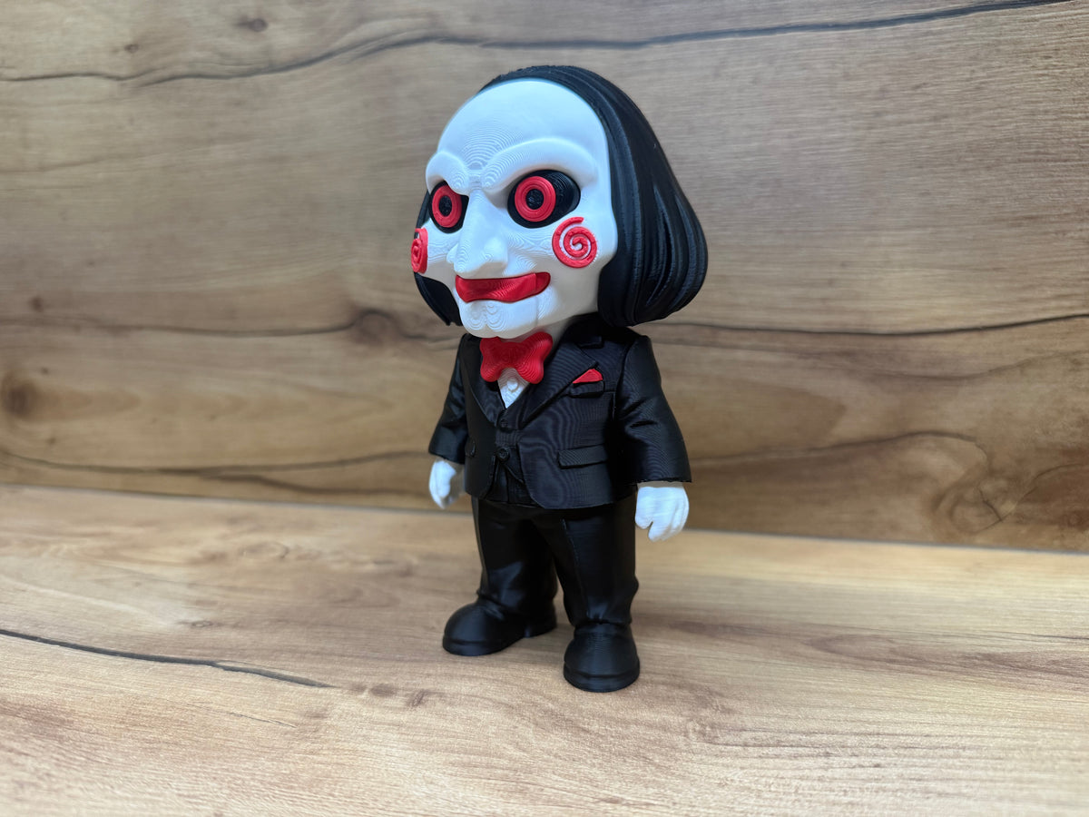 Billy the Puppet Saw Minifigure Sculpture — front three-quarter close view showing full face character accuracy including both red spiral cheek rosettes, red-ringed eyes, open teeth grin, forehead wrinkles, black bob hair and red bow tie against wooden background