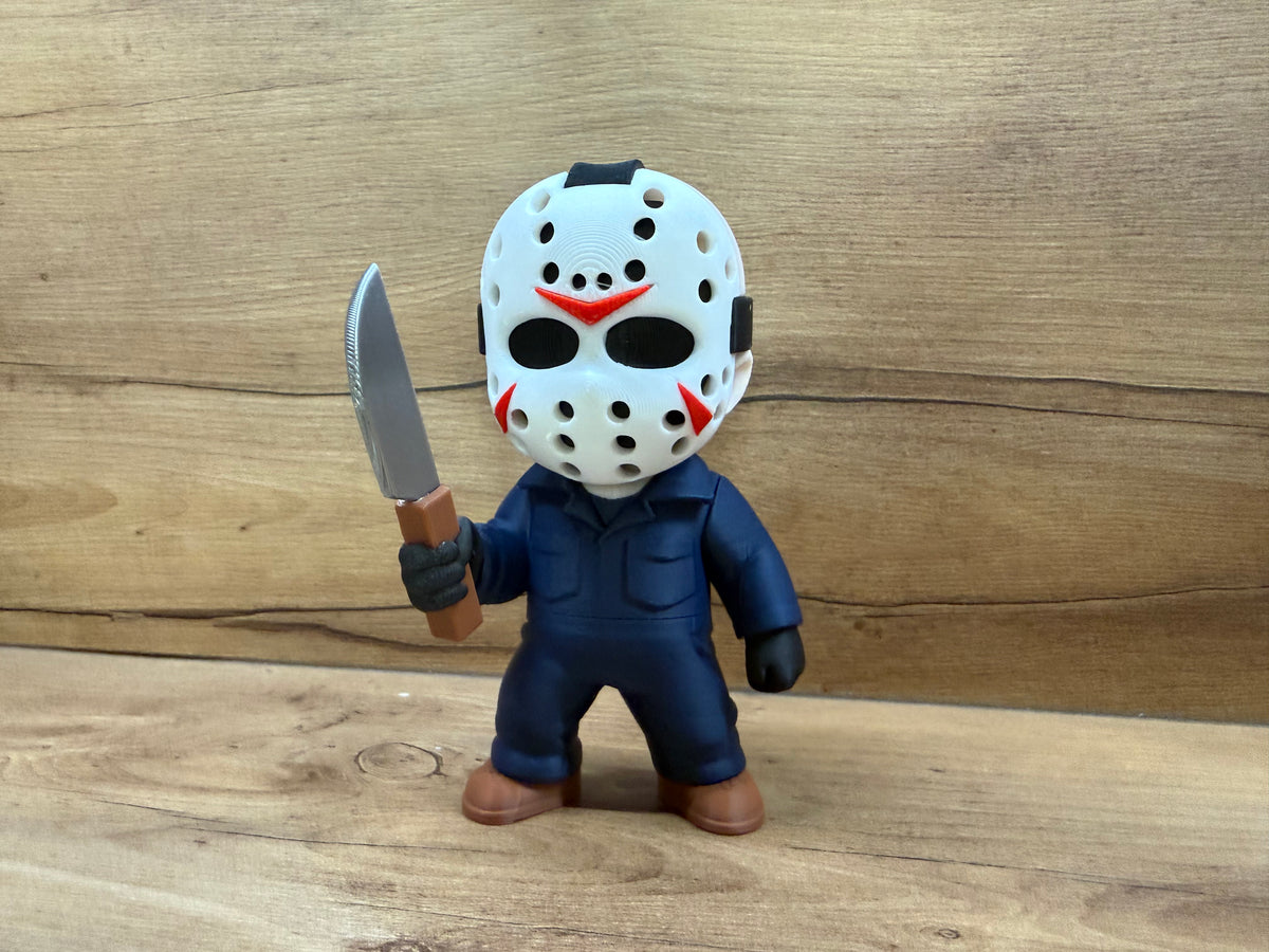 Jason Voorhees Friday the 13th Minifigure Sculpture — front view showing white hockey mask with black oval eye apertures, red chevron nose marking, red cheek accents, ventilation hole pattern and black crown strap, dark navy blue coverall with chest pocket, black gloves, right hand raised holding silver cleaver with brown handle, brown work boots