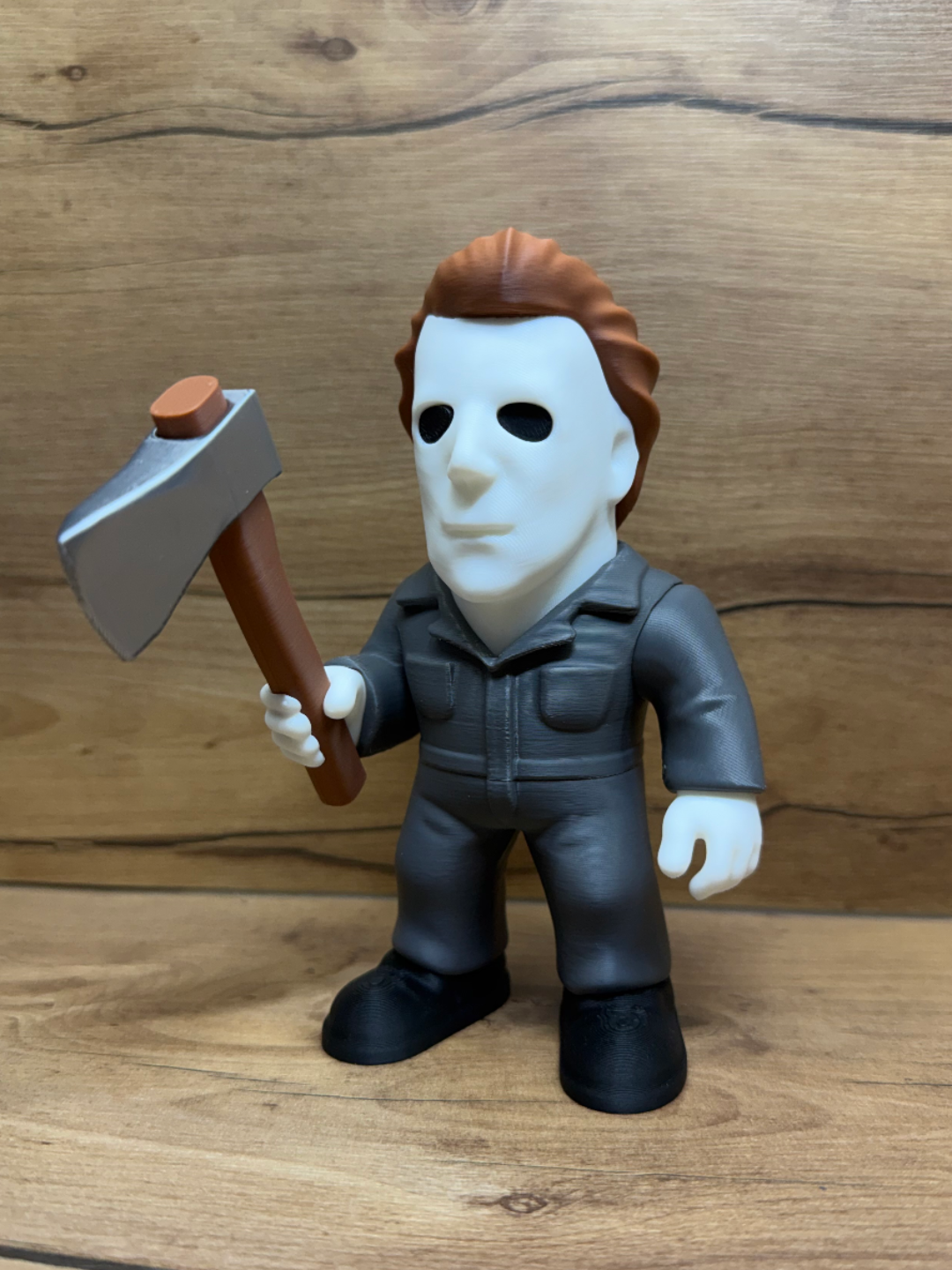 Michael Myers Halloween Minifigure Sculpture — front view showing white featureless mask with two deep black oval eye apertures and subtle nose ridge, warm auburn swept-back hair with individual strand texture, dark charcoal grey mechanic's coverall with left breast chest pocket, bare white hands, right hand raised holding large silver axe with brown wooden handle, black boots