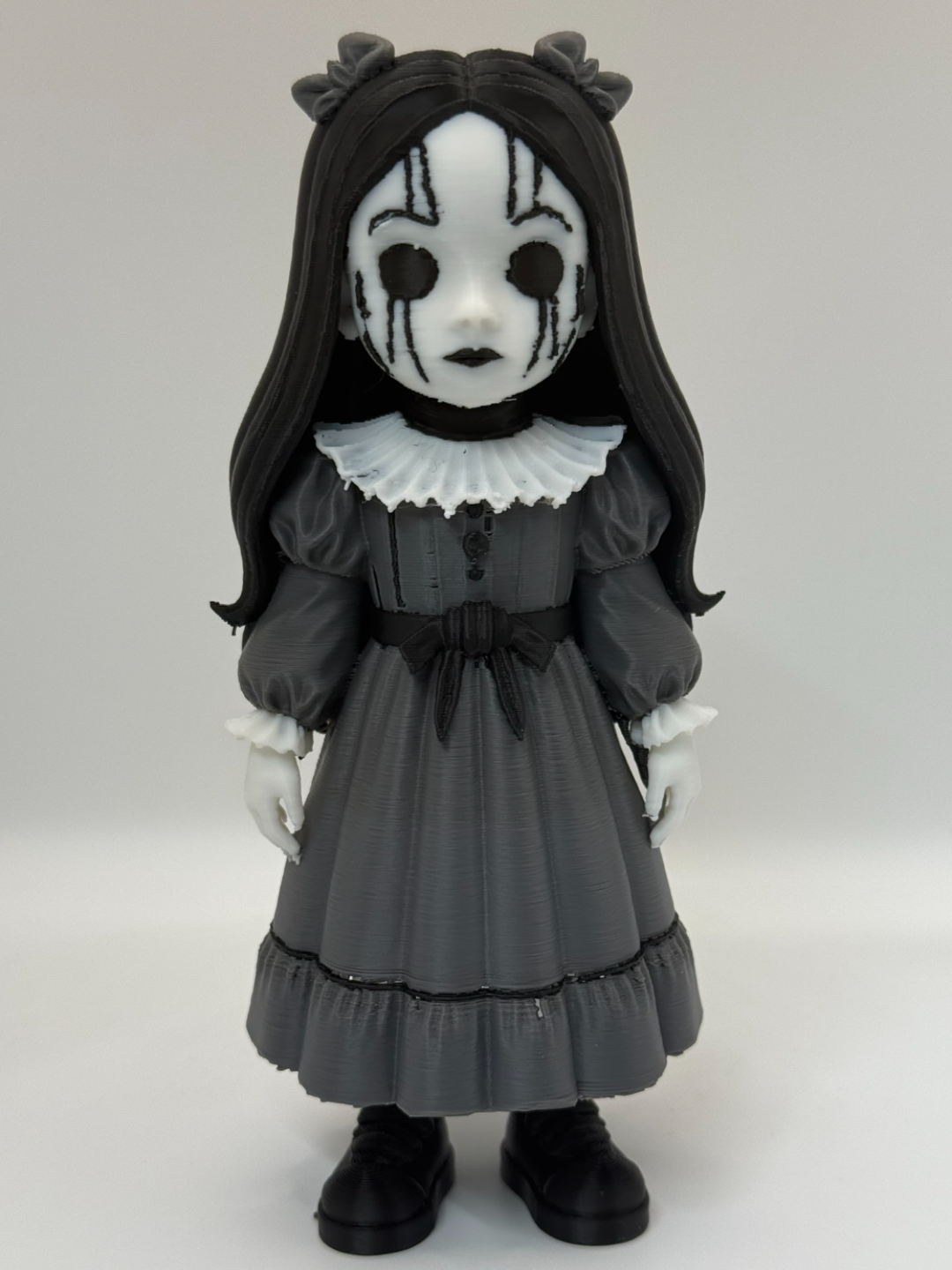 Gothic Doll Horror Minifigure Sculpture — front full body view showing white oval cracked mask with radiating black crack lines and four vertical drip lines below two large circular void eye apertures, small black-lipped closed mouth, long straight black hair with horn bow ornaments at crown, white Elizabethan ruffled collar, charcoal grey Victorian dress with three front buttons and black waist bow, white lace cuffs, double-tiered skirt with black trim and small black block-toe shoes