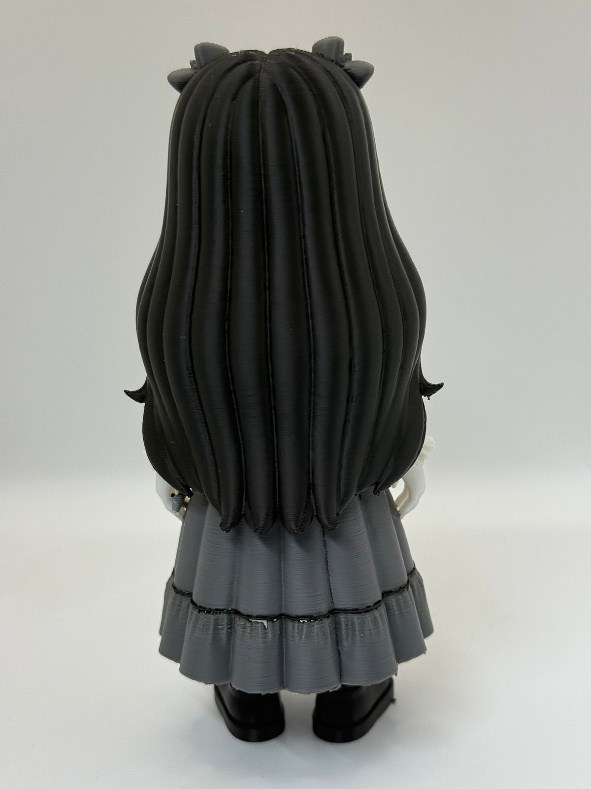 Gothic Doll Horror Minifigure Sculpture — rear full body view showing full black hair mass with deep individual strand texture from centre crown parting, small dark horn bow ornaments at crown top, charcoal grey Victorian dress rear with double-tiered skirt and black trim, white hands relaxed at sides and black block-toe shoes