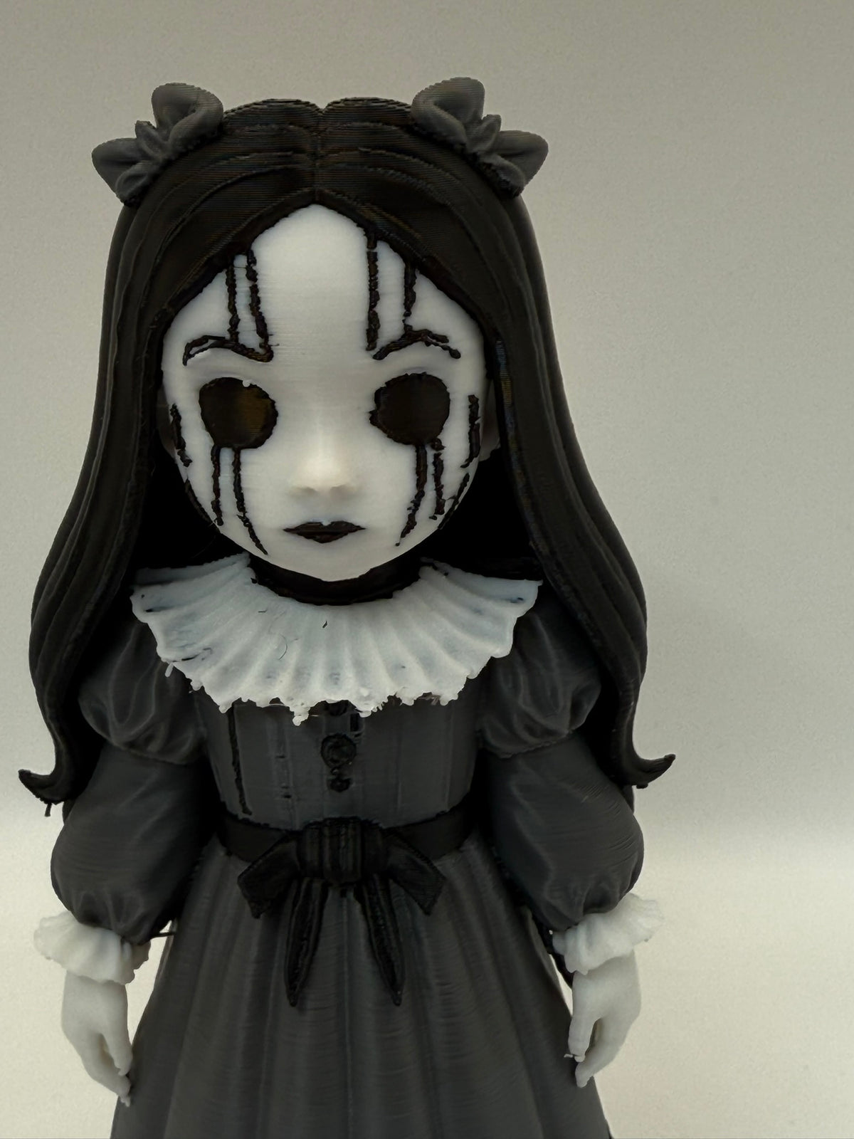 Gothic Doll Horror Minifigure Sculpture — front face close-up showing full white oval mask with radiating black crack lines across upper face, four vertical black drip lines descending from void eye aperture rims down cheeks, two large circular pure black void eye apertures with defined rims, small black-lipped closed mouth, white Elizabethan collar ruffle pleat detail and black hair framing the face on both sides with horn ornaments visible at crown