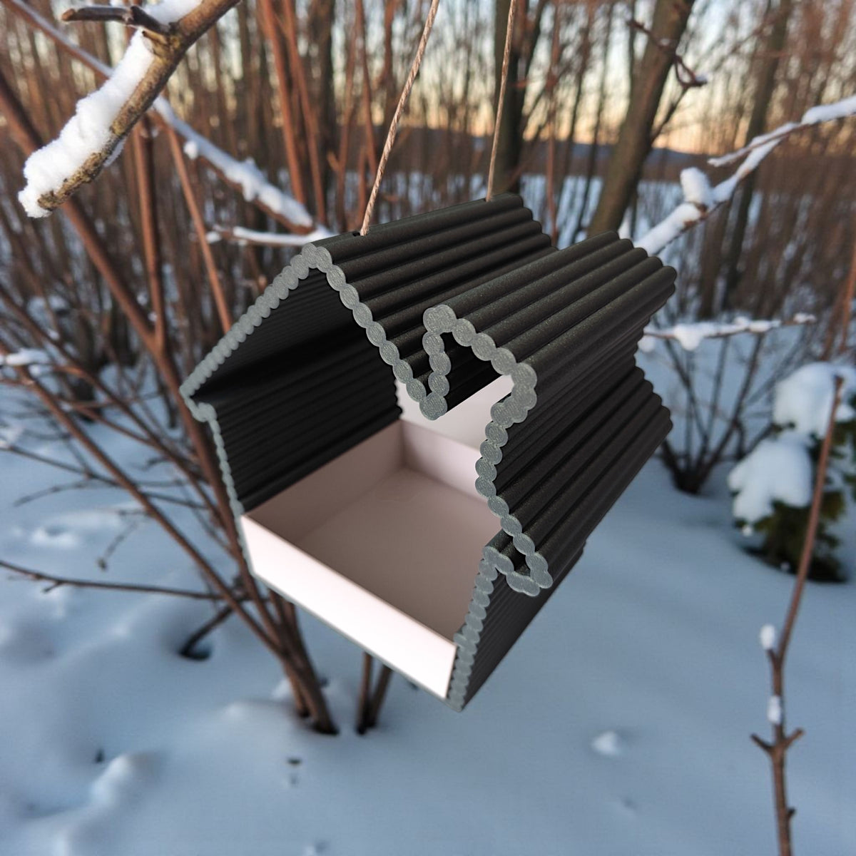 Black ribbed bird feeder hanging in snowy garden at sunset
