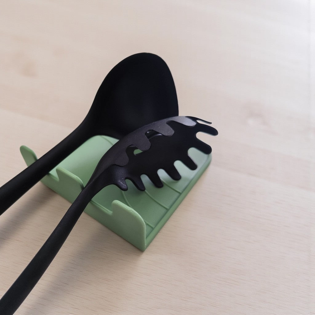 CookSitter utensil rest in green holding wooden spoon beside frying pan on induction hob, fresh tomatoes and basil nearby