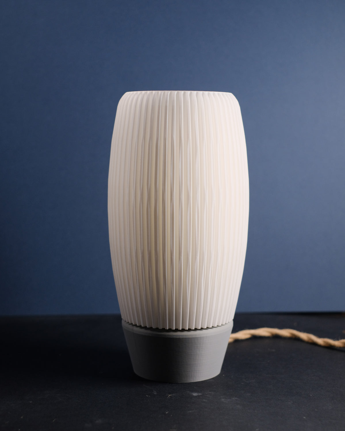 Ribbed E27 Lamp with diffuser tall oval barrel form dense fine vertical ribbing outer shade cream white matte finish grey rounded base warm inner diffuser glow deep navy blue background slight angle editorial hero shot