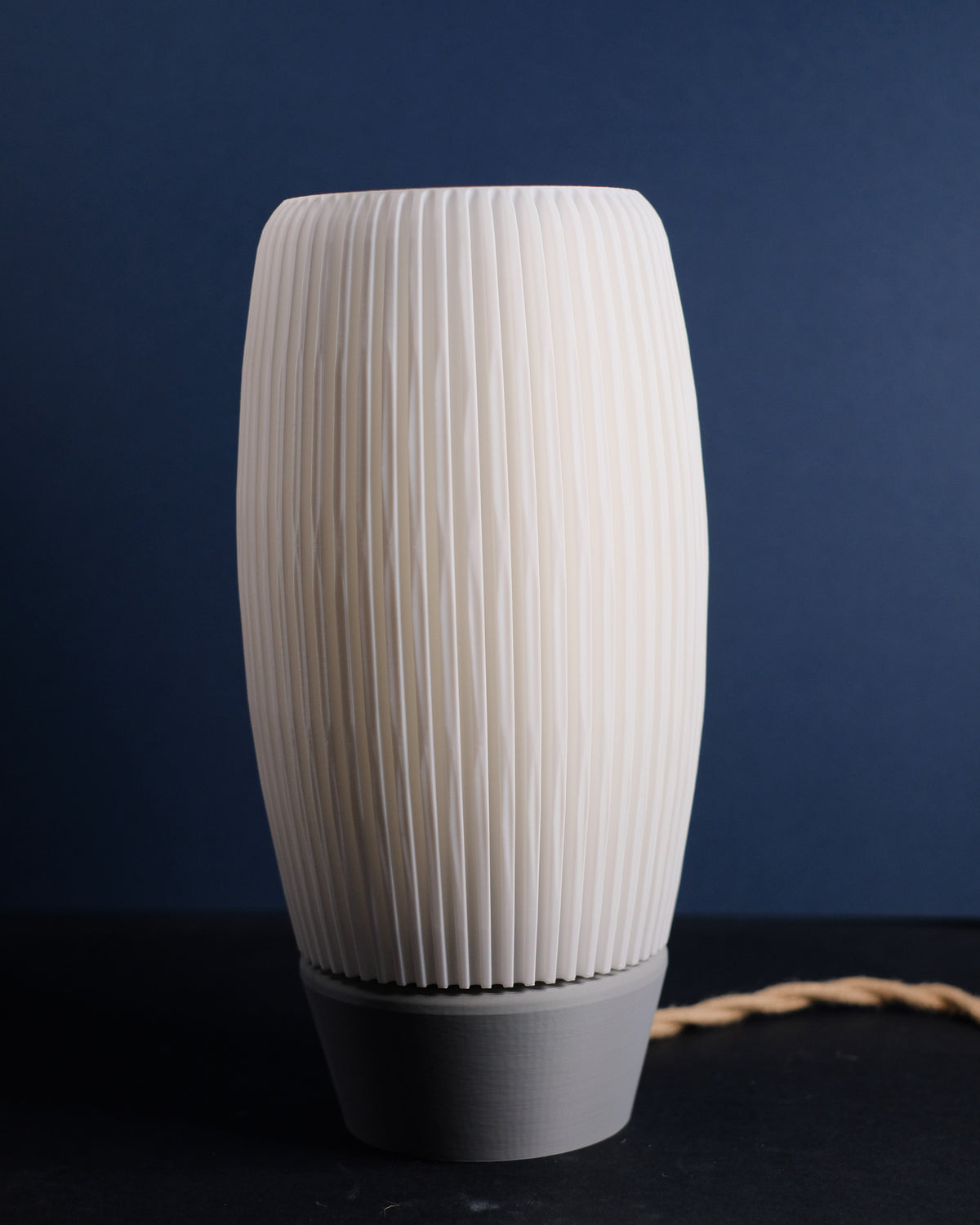 Ribbed E27 Lamp with diffuser tall oval barrel form dense fine vertical ribbing outer shade cream white matte finish grey rounded base warm inner diffuser glow deep navy blue background editorial hero shot
