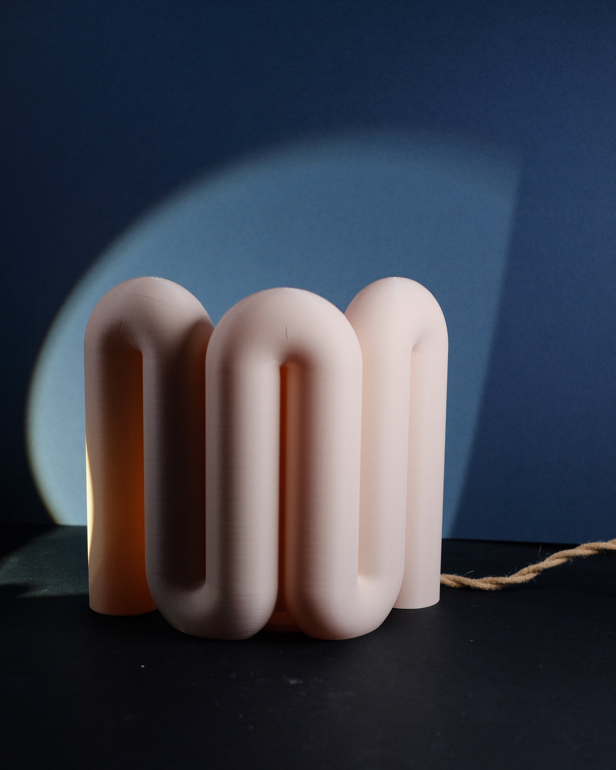 Wibble Curtain E27 Lamp compact square cluster four rounded arch tube loops warm amber light glowing through translucent blush pink matte body dark navy blue wall circular light arc behind deep blue background atmospheric editorial hero shot