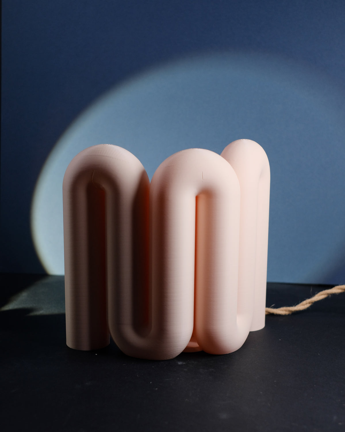 Wibble Curtain E27 Lamp compact square cluster four rounded arch tube loops warm amber light glowing through translucent blush pink matte body dark navy blue wall circular light arc behind editorial close angle hero shot