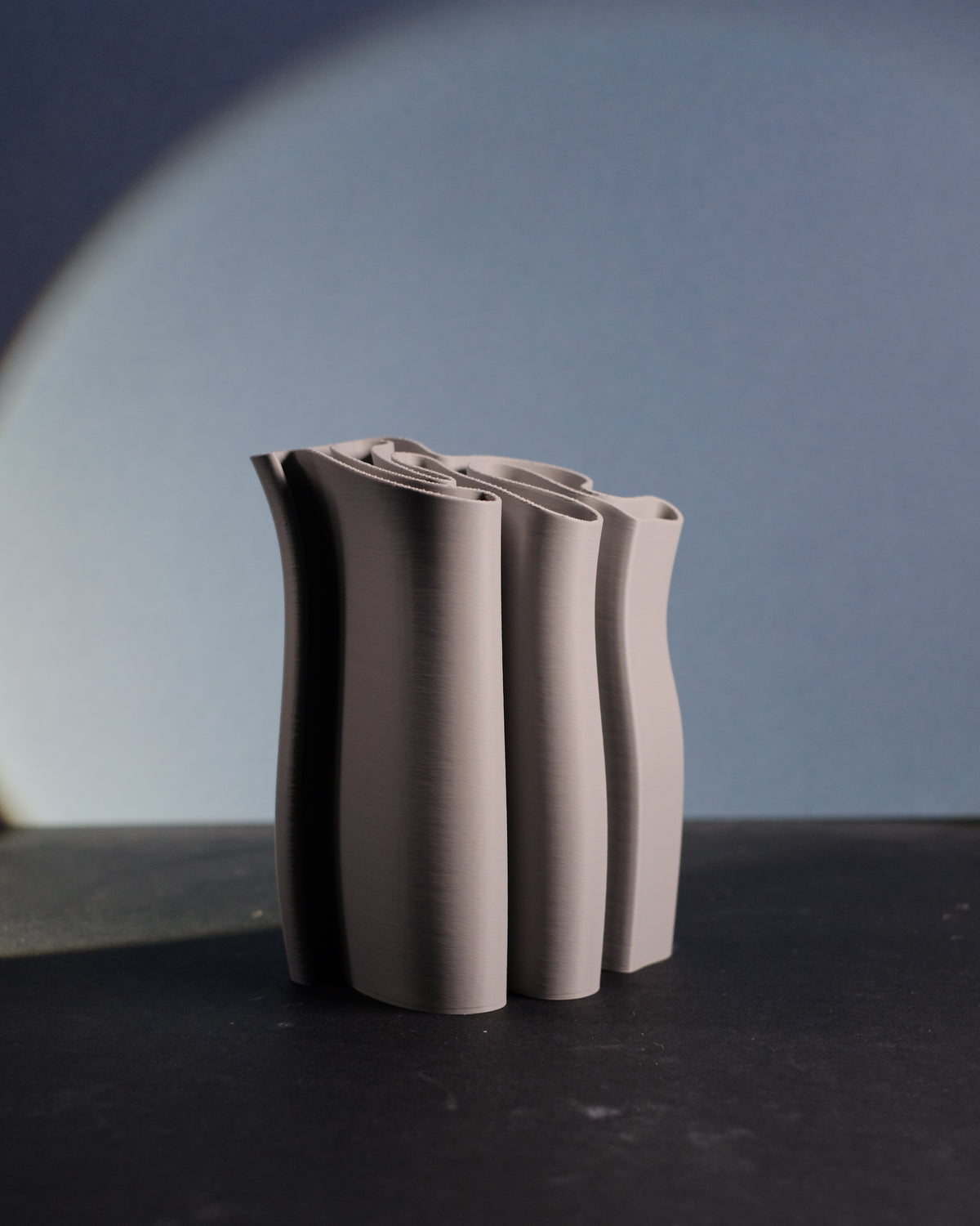 Fabric Ripple Vase and Desktop Organizer tall cluster undulating fabric-fold tube forms sinuous wavy ripple rim warm taupe matte finish blue-grey arc backdrop minimal surface editorial hero shot