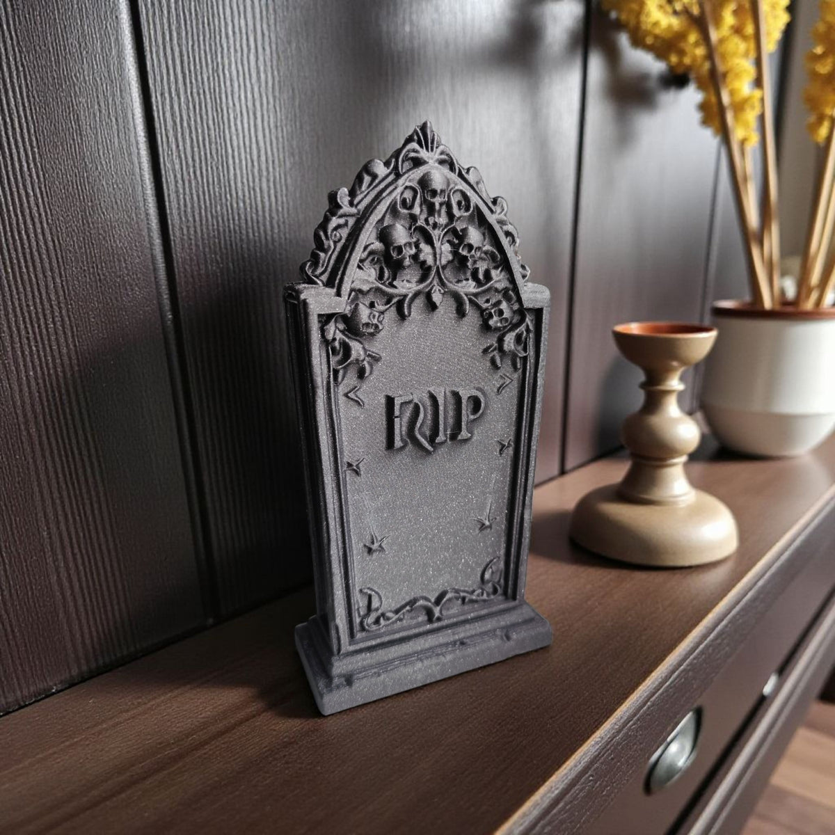 Scary Halloween decoration tombstone dark grey sparkle matte finish full figure on dark wooden sideboard showing deeply sculpted skull vine arch pointed gothic spire top raised RIP lettering star motifs moulded stepped base Halloween home decor scene