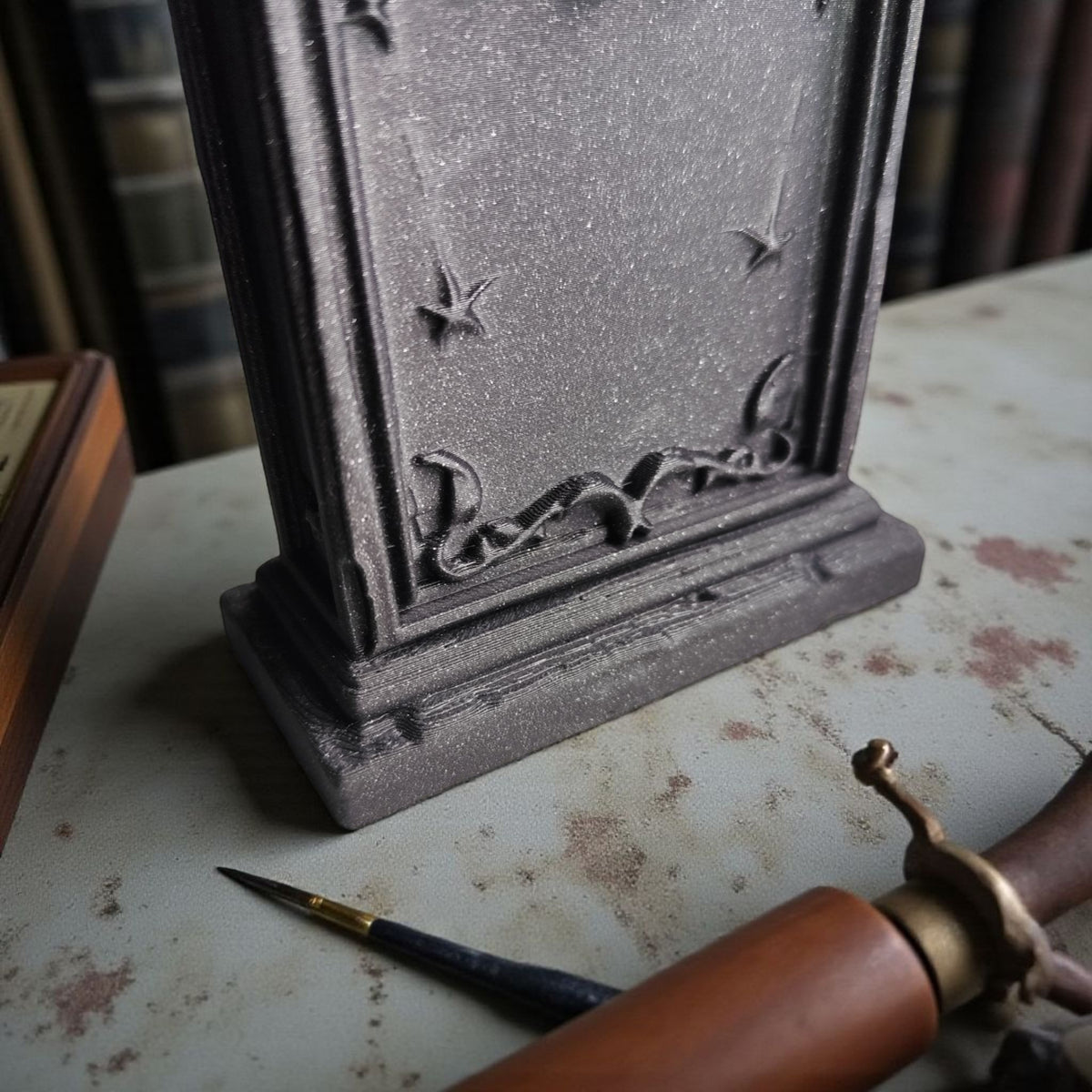 Scary Halloween decoration tombstone dark grey sparkle matte finish close up lower body and base detail deeply sculpted vine botanical scroll relief raised star motifs on aged pale surface antique brass quill pen atmospheric dark library editorial