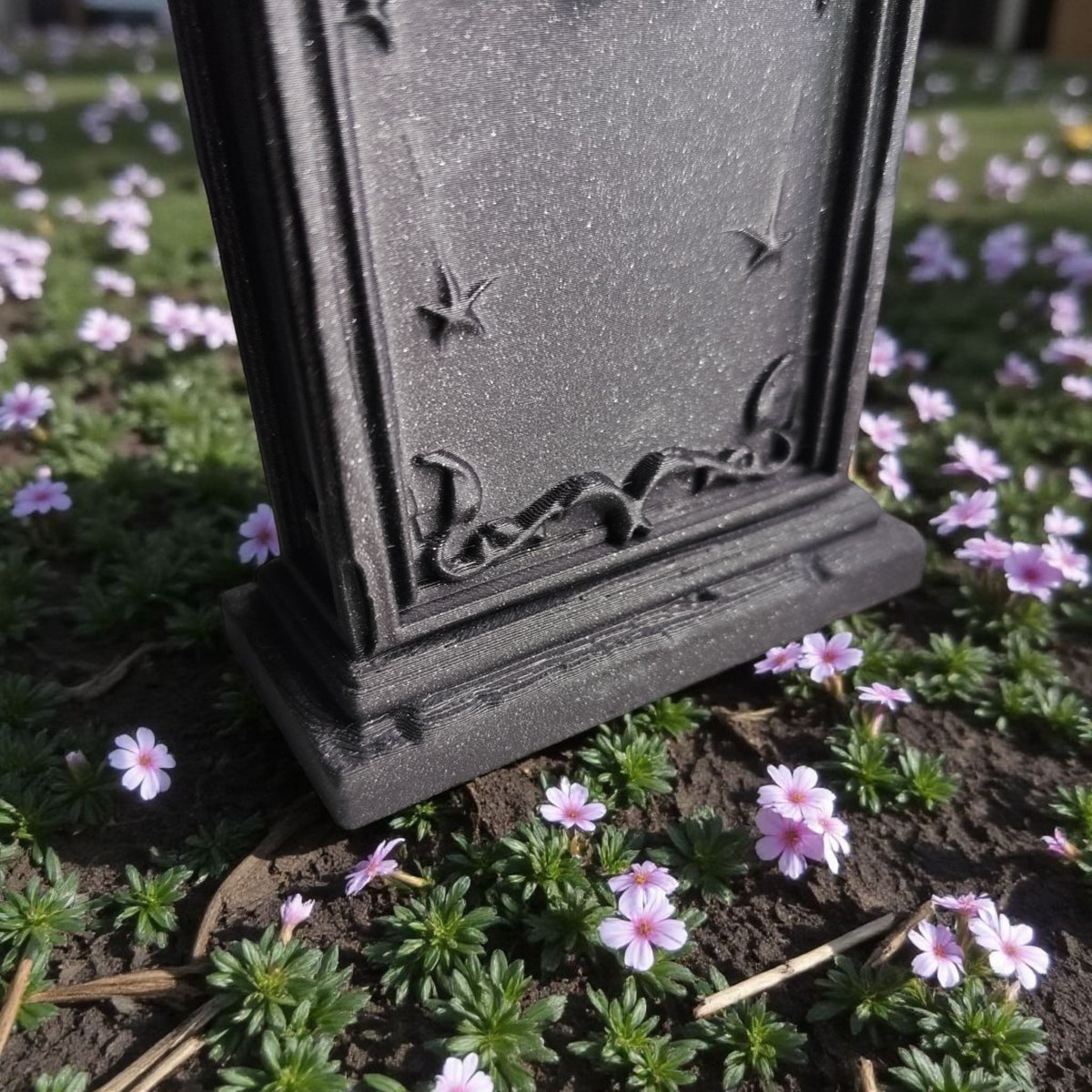 Grey gothic tombstone ornament base detail on ground with small pink flowers lifestyle