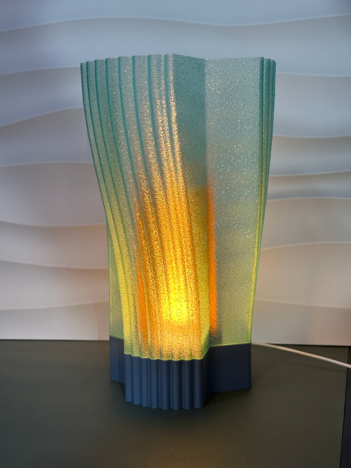 Swirl and Shine Desk Lamp real photo on draped concrete plinth dark background warm inner glow casting light through ribbed twisted shade