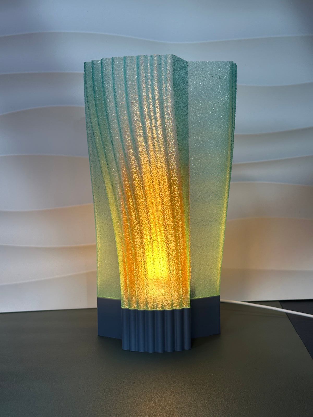 Swirl and Shine Desk Lamp real photo three quarter view on concrete plinth dark background warm glow through twisted ribs