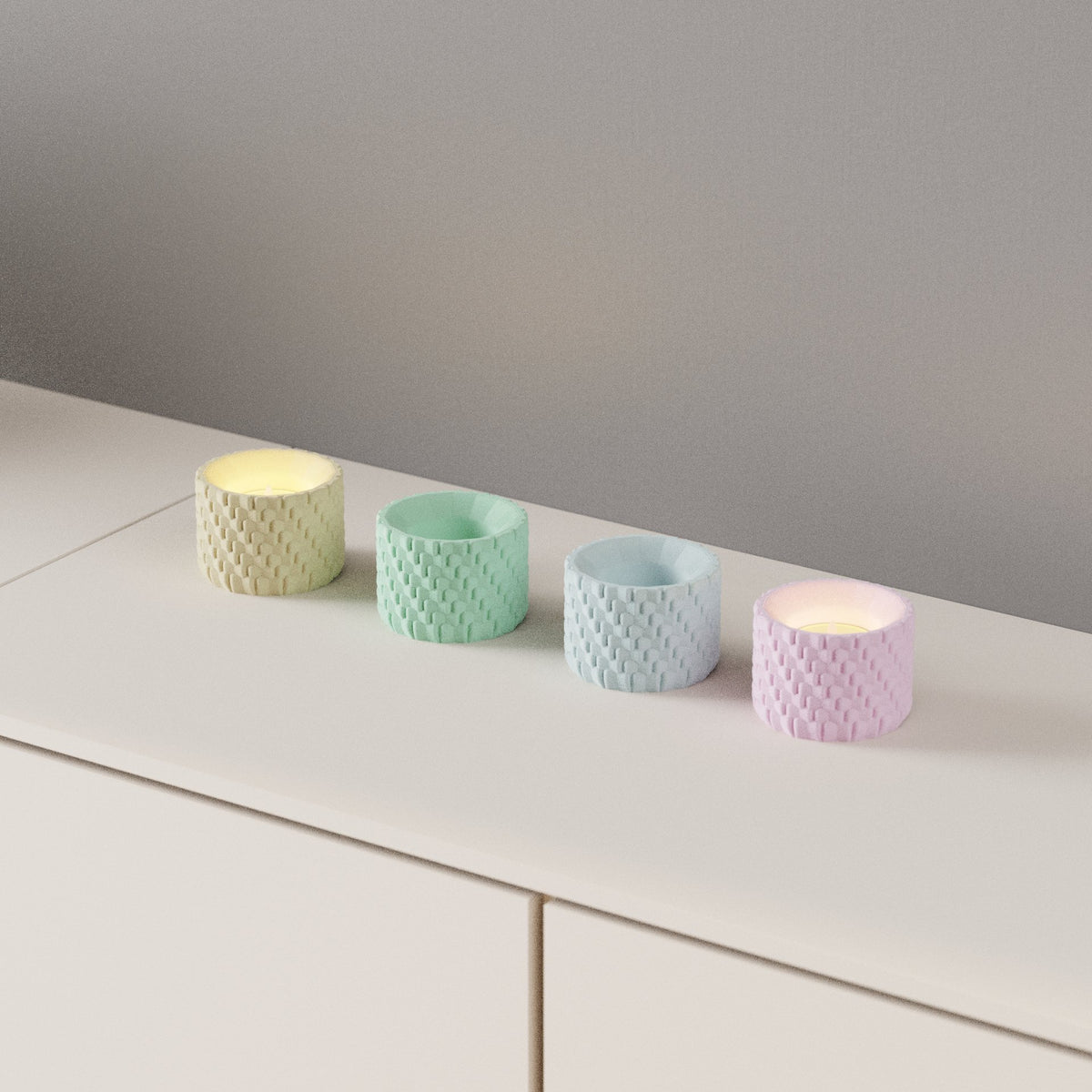 Woven Texture Tealight Holder four holders lemon yellow sage green powder blue lavender pink woven basket-weave surface glowing LED tealights pale sideboard wide shot