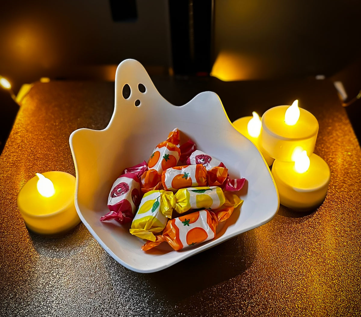White ghost-shaped sweet tray filled with wrapped treats beside warm lights