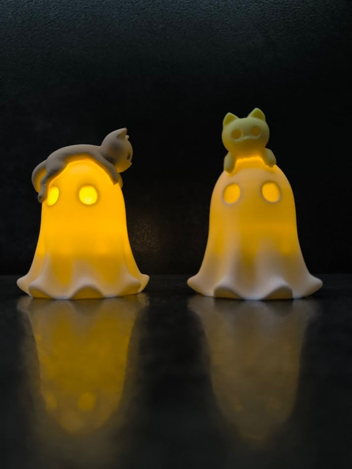 Two glowing white ghost tealight holders with cats, one cat curled across a ghost and one perched upright on the other
