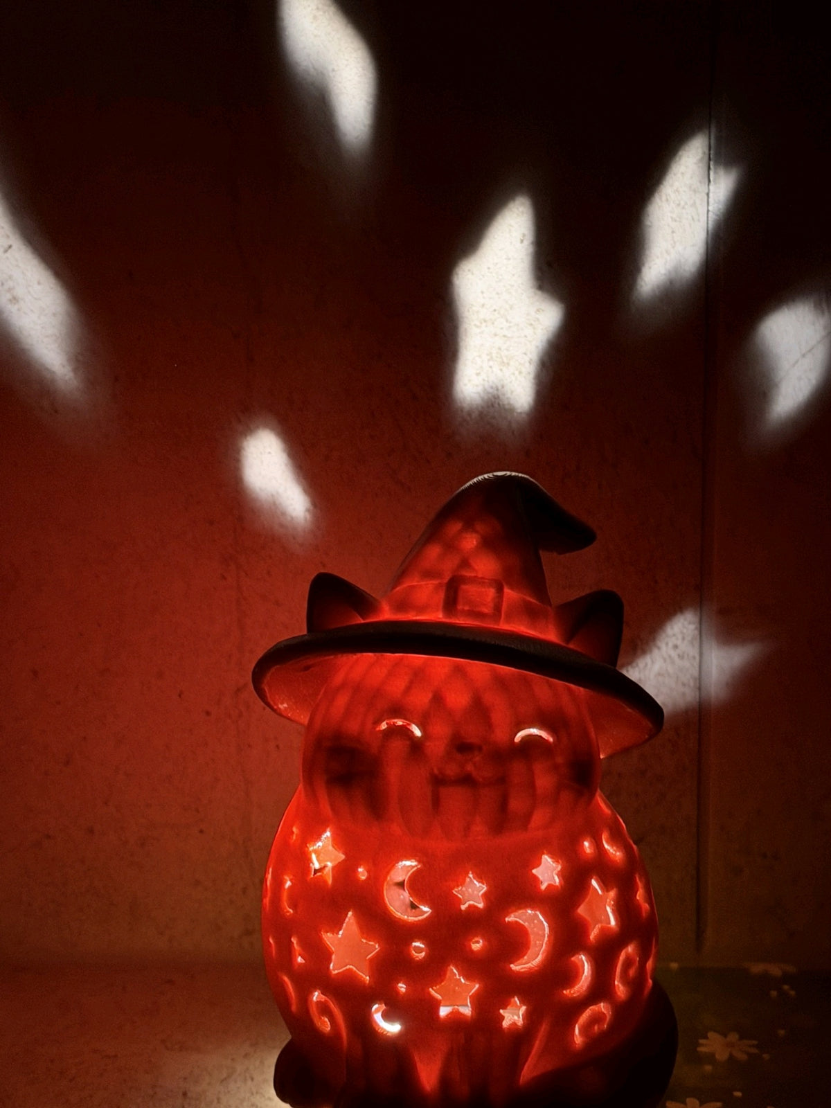 Orange witch-hat cat lantern glowing in a dark room, casting moon and star shapes onto the wall
