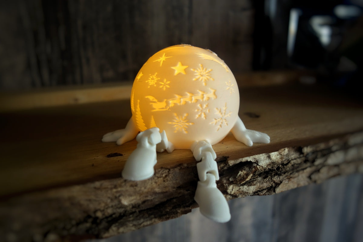White spherical tealight holder with a festive train detail and short sculpted legs, shown on a rustic wooden ledge