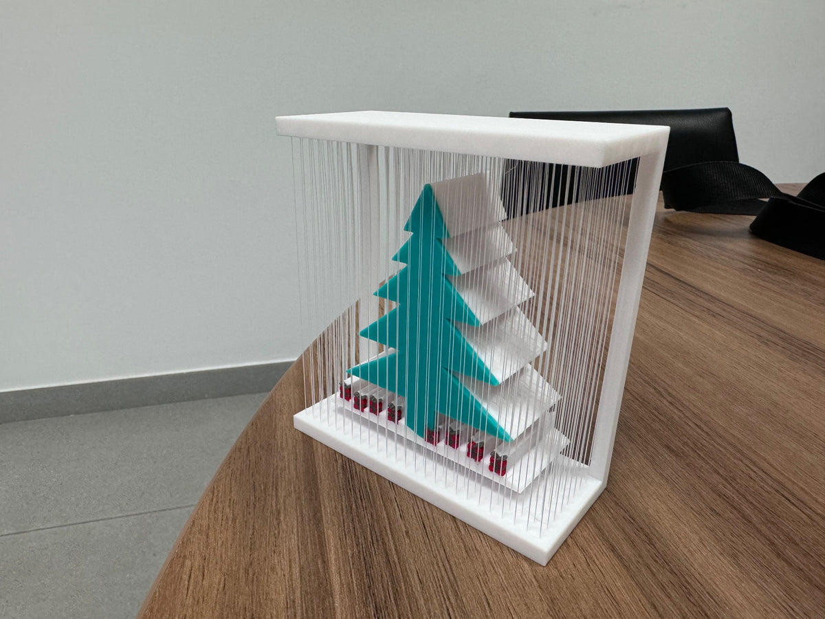 White rectangular frame with vertical strands surrounding a teal-green Christmas tree and red-and-dark base details, viewed on a wooden table.