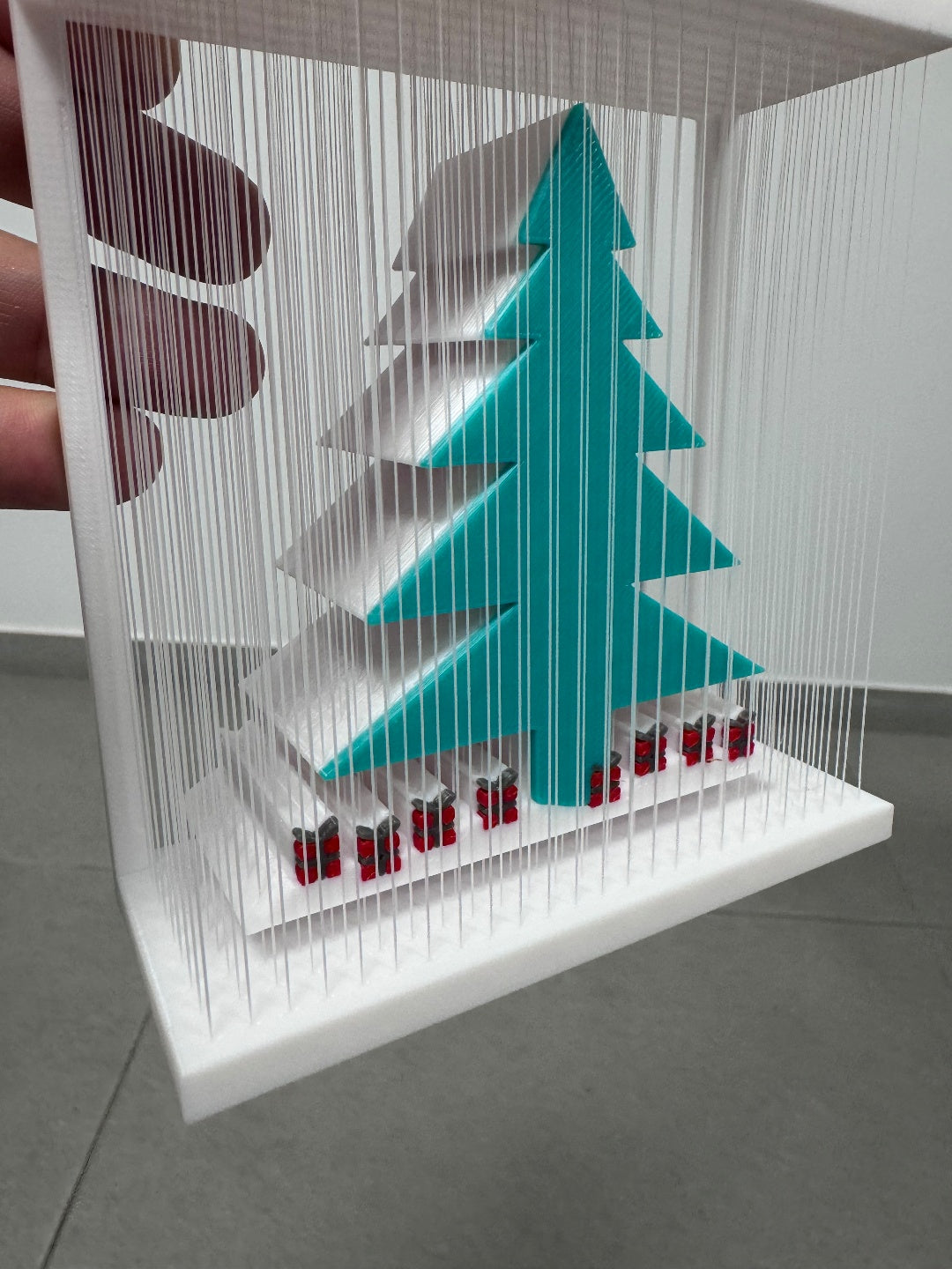 Close-up of the teal-green Christmas tree behind white vertical strands, showing the small red-and-dark details at the base.
