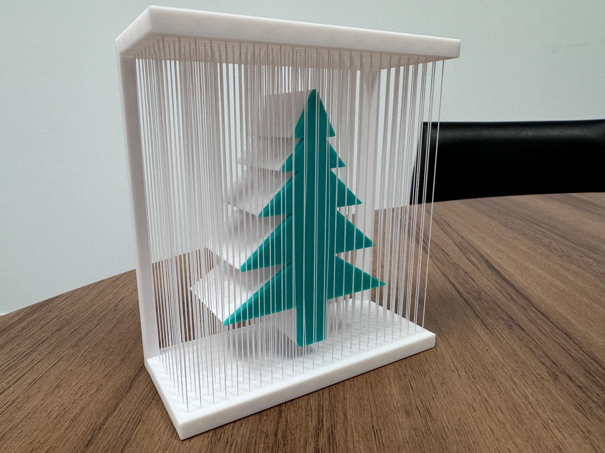 Teal-green Christmas tree inside a white rectangular frame with fine vertical strands, shown on a wooden table.