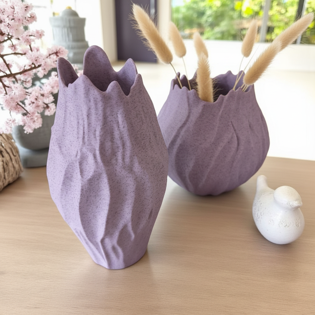 Two organic stone-like sculptural vases in dusty purple on a wooden side table with pampas grass