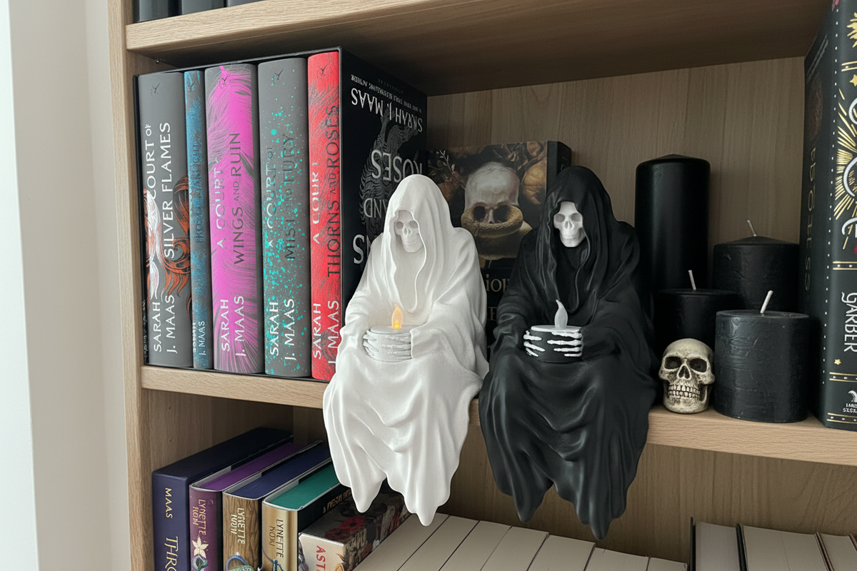 Two Grim Reaper shelf sitter figurines white and black hooded skull faces flowing robes skeletal hands holding glowing candles dark wooden bookshelf colourful hardcover books black candles Halloween shelfie landscape editorial